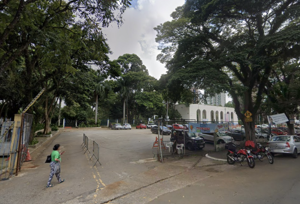 EMBARQUE - PARQUE CERET - Área externa arborizada, com estacionamento asfaltado. À esquerda, uma mulher caminha próximo a um portão gradeado. Ao centro, prédio branco com arcos, cercado por carros, uma van escura e motos estacionados. Ao fundo, prédios altos entre as árvores.