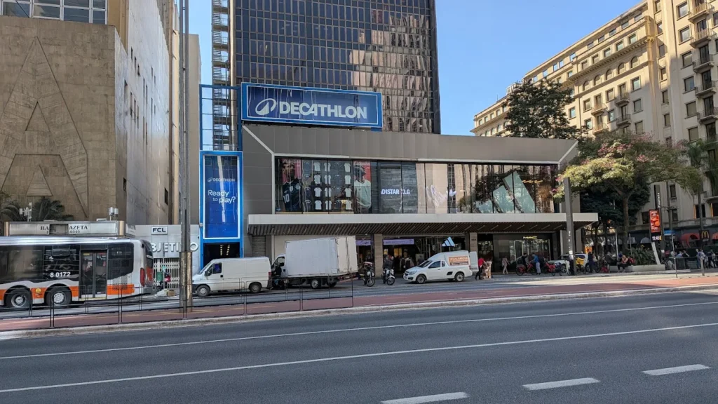 LOJA DECATHLON - Prédio comercial com fachada de vidro em avenida larga. Na parte superior, letreiro azul retangular escrito “Decathlon” em letras brancas. Nas vitrines, há roupas e produtos esportivos. Na calçada, há faixa de pedestre, um ônibus, uma van e um caminhão estacionados, e pessoas caminhando. Ao fundo, prédios altos, um deles com fachada espelhada.